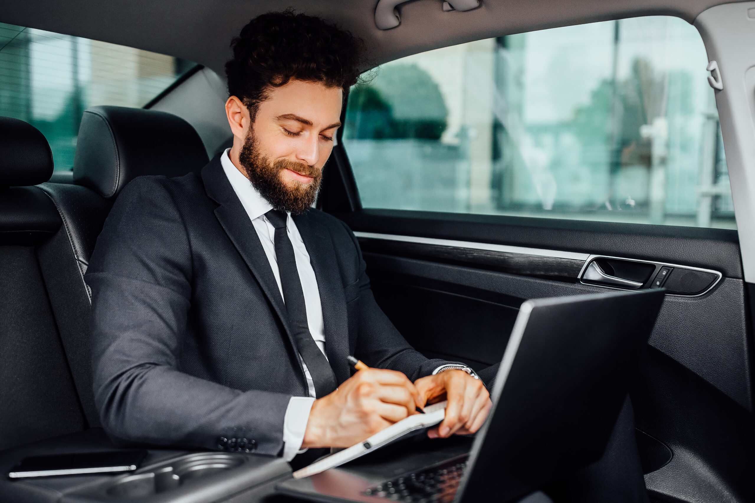 handsome bearded smiling businessman working backseat car makes notes notebook from his laptop 1 scaled 1